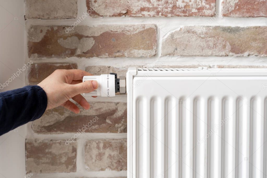 Cropped shot of woman shot adjusting temperature on white radiator with thermostat against brick wall background in loft style room, person using central heating system at home