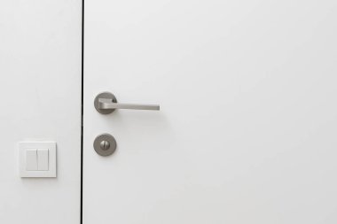 Kapalı beyaz kapılar ardında güvenlik kilidi olan açık gri renkli basit metal kapı kolu ve otel odasındaki beyaz duvardaki ışık düğmesi.