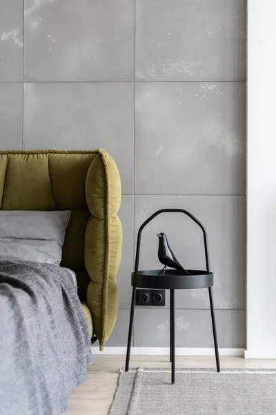 Vertical shot of minimal style bedroom in monochrome colors with big comfy bed covered with gray bedding next to industrial metal coffee table decorated with artificial black bird on carpeted floor
