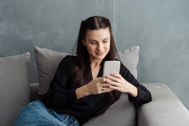 Genç gülümseyen bir kadın modern akıllı telefonunda online uygulama kullanıyor, mesaj gönderiyor, blog okuyor ve rahat bir oturma odasında rahat bir koltukta oturuyor. Kadın evde dinleniyor ve internet sitesinde sörf yapıyor