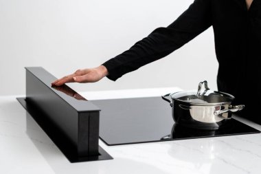 Cropped shot of female adjusting built in induction ceramic cooktops exhaust, metal pot with food on stove, cooking in minimalistic kitchen with glossy white counter and integrated appliances