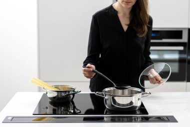 Cropped shot of woman boiling water in metal pot to cook spaghetti, holding glass lid and big spoon while cooking on black ceramic induction cooktop in white minimalistic kitchen
