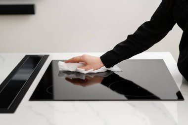 Woman cleaning black reflective glass induction stove with white rag, making surface shiny and clean, washing cooktop after cooking food on it in simple black and white themed kitchen, cropped shot