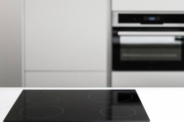 White countertop with glossy built in ceramic black induction stove, big white cupboard with built-in electrical oven in empty kitchen with nobody in sight, blurred background