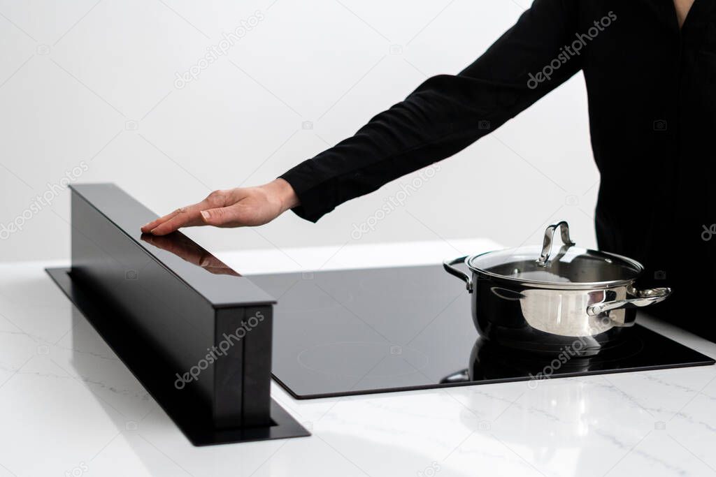 Cropped shot of female adjusting built in induction ceramic cooktops exhaust, metal pot with food on stove, cooking in minimalistic kitchen with glossy white counter and integrated appliances