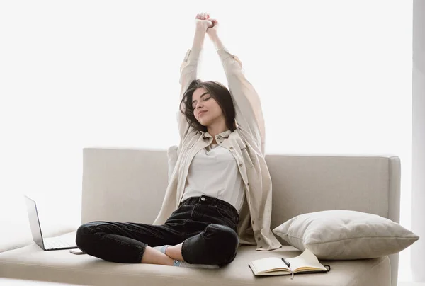 Full length view of dreamy and serene young woman sitting on comfort couch in living room, resting at home, raised hands up after work on laptop computer in home office