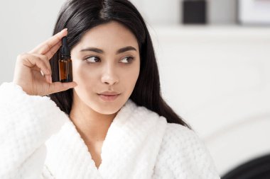 Doğal güzellik ve organik kozmetik reklamları. Suratı kırışıklık önleyici serumla dolu şişeyi tutan Asyalı genç bir kadının portre resmi, yan bakıyor, fotokopi uzayının beyaz arka planında duruyor.