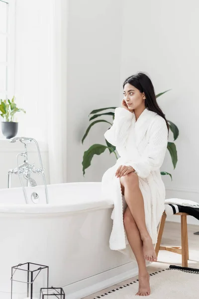 自然美概念 年轻的亚洲女人穿着柔软的浴衣坐在白色浴池上 望着旁边的垂直镜头 妇女在现代化的室内浴室度过上午 — 图库照片