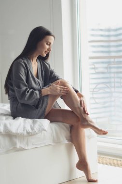 Esmer genç bir kadın yatak odasında kıllarını aldırdıktan sonra ipek bacaklarına nemlendirici krem sürüyor. Kişisel bakım günü, hijyen ve sağlık konsepti