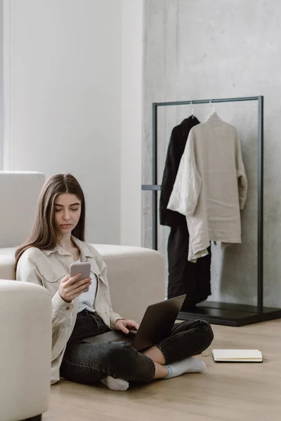 Online education and internet app concept. Vertical view of young woman in casual clothes looking at smartphone, working with laptop at home office, sitting on floor near comfort couch in living room