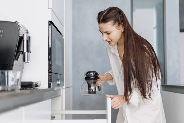 Young woman with long brunette hair in homewear taking tea pot from cupboard, preparing hot morning drink while standing in stylish fully furnished kitchen at home