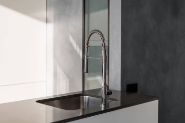 Parlak mutfak tezgahı, uzun, metalik, parlak musluk ve kare şeklinde lavabo, basit tasarımla iç mekan, pencereden gelen ışıkla aydınlatılmış oda.