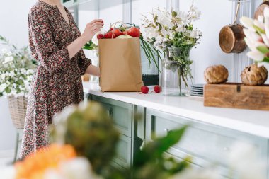 Elbise giymiş bir kadın market poşetindeki sebzeleri açıyor, beyaz yüzeyi olan eski yeşil bir tezgaha koyuyor, çiçek ve yeşilliklerle dolu rahat bir mutfak ortamında duruyor. Kırpılmış atış