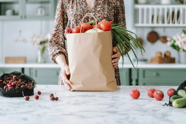 Woman wearing dress holding paper grocery bag and resting it on kitchen island with shiny marble surface, filled with healthy vegetables, green furniture in blurred background. Cropped shot