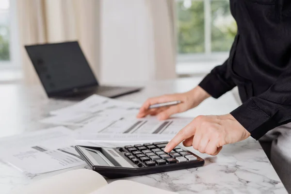 Person in black shirt calculating monthly bills and debts with help of calculator and laptop, invoices laying around on marble table with windows in blurred background. Cropped shot