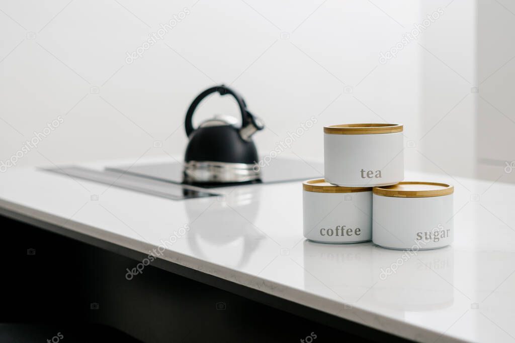 Black whistle kettle being boiled on glass electric stove with three stacked labeled storage canisters in front of it. Jars with coffee, tea and sugar on white table in kitchen. Blurred background