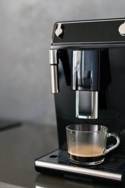 Kahve makinası bittikten sonra espresso hazır ve bekliyor köpüklü sıcak içecek, metalik parçalı siyah ev aleti, temiz mobilya, bulanık arka plan. Dikey çekim