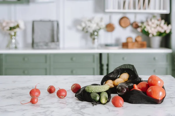 Various vegetables laying in black mesh bags on kitchen island with marble surface, tomatoes, potatoes and radishes, green counter with lots of flowers in blurred background