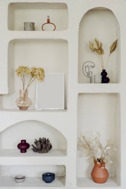 Duvar dekorunun dikey görüntüsü, apartmanın içindeki beyaz raflar. Ev için klasik dekorasyon, İskandinav odasındaki oyukların kapatılması. Vazoda kuru bitkiler olan doğal dekor..