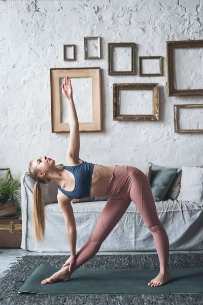 Vertical shot of beautiful fit woman in good shape doing morning gymnastics while standing in yoga asana Utthita Trikonasana in living room at home, sporty female in active wear exercising indoors