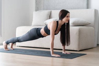 Esmer, genç bir kadın egzersiz yapıyor, karın kaslarını çalıştırmak için tahta pozisyonda duruyor, kollarına dayanıklılık ve oturma odasında yoga minderinin üzerinde dayanıklılık. Sağlıklı yaşam tarzı, spor ve spor konsepti