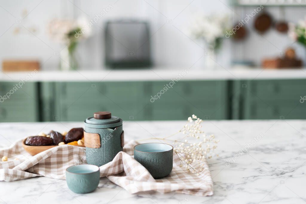 Marble kitchen island, teal teapot set with couple small cups next to bowl filled with dried fruits and nuts laying on tablecloth, green counter with drawers and white top in blurred background