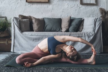 Güzel, sportif genç bir kadın yoga yapıyor ve ev antrenmanı sırasında dizlerinin üstüne çöküp egzersiz yapıyor. Parivrtta Janu Sirsasana 'da oturan bir kadın oturma odasındaki spor minderinde poz veriyor.