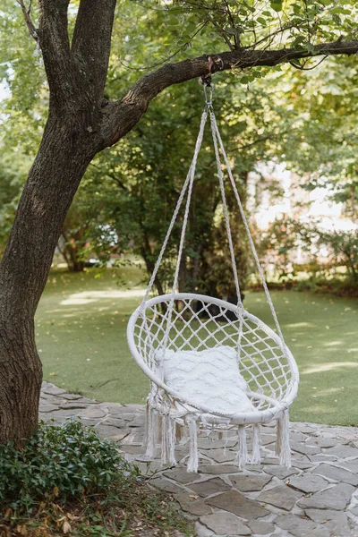 Swing furniture with nobody, wicker chair design. Outdoor white boho armchair for summer relax at nature, garden decoration with seat. Chair hanging at tree for comfortable leisure.