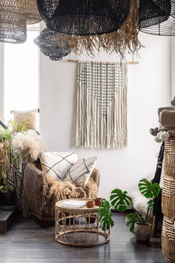 Boho şıklığında, rahat bir oturma odası. Ev dekorunun dikey görüntüsü, doğal malzeme mobilyaları, yastıklı koltuk, bambu kahve masası ve hasır avizenin altındaki duvarda makrome.