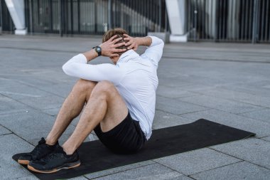 Adam yoga minderinde karın kası egzersizi yapıyor. Spor giyim sektöründe arka planda kalan sporcu, kenarda durup günlük egzersizlerle vücudunu güçlendiriyor.