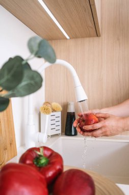 Kadın elleri, çağdaş bir mutfak ortamında, şeffaf, beyaz bir musluğun altında, dikey çekimde taze kırmızı elmayı yıkıyor. Sıcak ahşap tonlar ve dolapların minimalist tarzı.