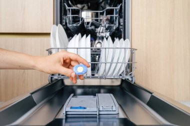 Bir el modern bulaşık makinesinin kompartımanına bulaşık makinesi koyar. Temiz tabaklar ve çatal bıçaklar üst rafta, yıkanmaya hazır..