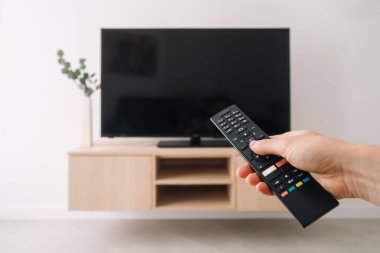 POV çalışanı uzaktan kumanda tutuyor, televizyon izlemeye hazır, evde eğlenmeye hazır. Ahşap bir dolaba monte edilmiş büyük düz ekran televizyonu olan şık bir oturma odası..