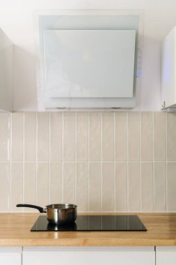 Modern beyaz mutfak kaputunun altındaki elektrikli bir ambarda metal bir tencere duruyor. Tahta tezgahta indüksiyon sobası, dikey çekim. Evde yemek pişirmek.