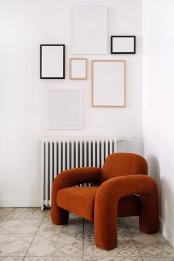 Boş çerçevelerle süslenmiş beyaz duvara karşı rahat turuncu koltuklu şık oturma odasının dikey görüntüsü. Ev dekorasyonu ısıtma radyatörünün üstünde