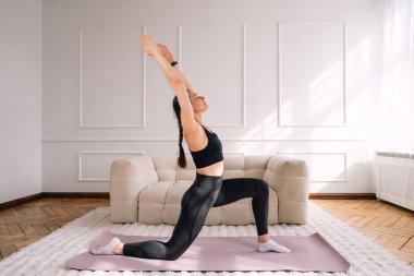Genç bir kadın oturma odasındaki yoga minderinde spor yapıyor. Parlak bir dairede uzanıyor, sağlıklı bir yaşam tarzı ve fiziksel aktivitesini teşvik ediyor. Yoga kavramları