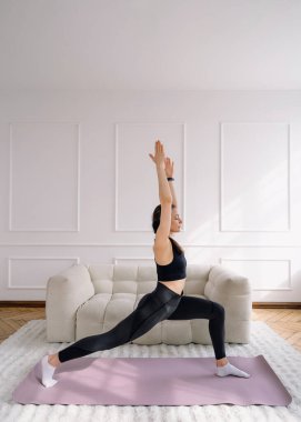 Genç bir kadının profil fotoğrafı yoga pozisyonunda dengesini oturma odasındaki bir paspasın üzerinde sağlıyor. Derin nefes alıyor, aktif giysiler giyiyor ve sağlıklı ve aktif bir yaşam tarzına odaklanıyor.