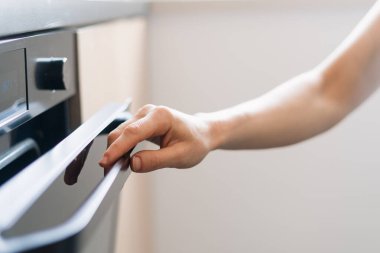 İyi tasarlanmış bir mutfakta modern elektrikli fırının kapısına uzanan bir el. Çağdaş ev aletleri ev ortamında verimli yemek yapmayı temsil eder..