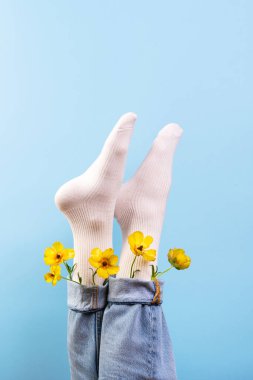 Parlak beyaz çoraplar içinde şen şakrak sarı çiçeklerle çevrili kot pantolonlardan dışarı bakarak açık mavi bir arka plana kurulmuş canlı ve neşeli bir atmosfer yaratıyor..