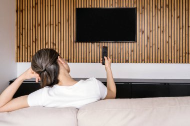 Öncü TV ekranı, model. Oturma odasındaki bir kanepede uzanan kadın, uzaktan kumandalı ahşap bir duvar paneline monte edilmiş akıllı bir televizyonu kontrol ediyor. Mekanda modern mobilyalar ve rahat bir atmosfer var..