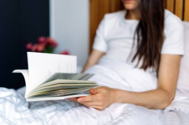 Şık bir otel odasında, misafir rahat bir yatakta rahatlar, kağıt sayfaları çevirir, kitap okur. Modern dekorlar ve yumuşak çarşaflarla çevrili bir kadın, sakin bir atmosfer yaratıyor.