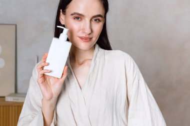 Cilt bakım ürününü banyoda gösteren genç bir kadın, rahat bir keten bornoz giyiyor. Sabah rutininin bir parçası olarak sağlığı ve sağlığı somutlaştırıyor. Ev spa 'sını şımartmaktan zevk alıyor..