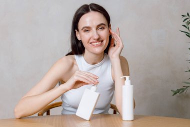 Genç bir kadın banyodaki ahşap bir masada oturuyor ve cilt bakımı ürünleri uyguluyor. Güzellik ayininde nemlendirici şişeler sergilerken, sağlığını ve özgüvenini somutlaştırırken gülümsüyor..