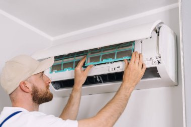 Bir teknisyen, bir apartman dairesinin duvarına monte edilmiş modern bir klimanın hava filtresini değiştiriyor. Hizmet, konforlu yaşam için ev aletlerini güvence altına alır.