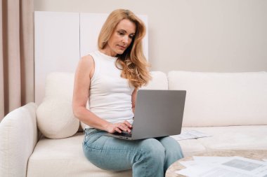Female focused on her laptop, working remotely online at home. The workspace is elegant and minimalistic. A mature woman using pc computer sits comfortably on a sofa in her modern apartment.