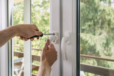 Bir tamirci elinde tornavida aleti tutuyor, plastik bir kabzayı yerleştiriyor ve bir apartman dairesine yeni bir pvc pencere çerçevesi yerleştiriyor. Kırpılmış insan eli.