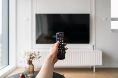 Smart TV is mounted on the wall in a contemporary living room while a person holds a remote control change channels, ready to watch movie. Entertainment options in modern home.