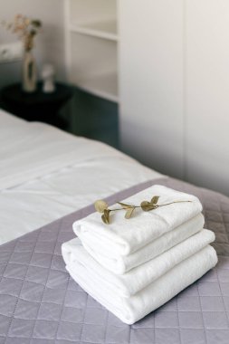 Düzenli bir otel odası, modern yatak takımlarıyla düzgünce yapılmış bir yatak. Taze beyaz havlular zarif bir şekilde yatağın üzerine istiflenir, üzerlerinde dekoratif bir dalla tamamlanır..