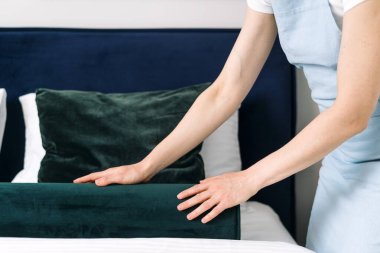 Bir insan, modern bir otel odasında düzenli yapılmış bir yatağa dekoratif bir yastık yerleştirir. Zarif bir tasarım sergiler ve misafirler için davetkar bir atmosfer sunar..