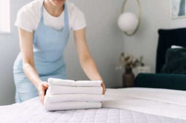 Bir otel çalışanı, çağdaş bir yatak odasında temiz beyaz havluları düzgünce yapılmış bir yatağa dikkatlice diziyor. Odada şık bir dekor ve rahat bir ortam var..